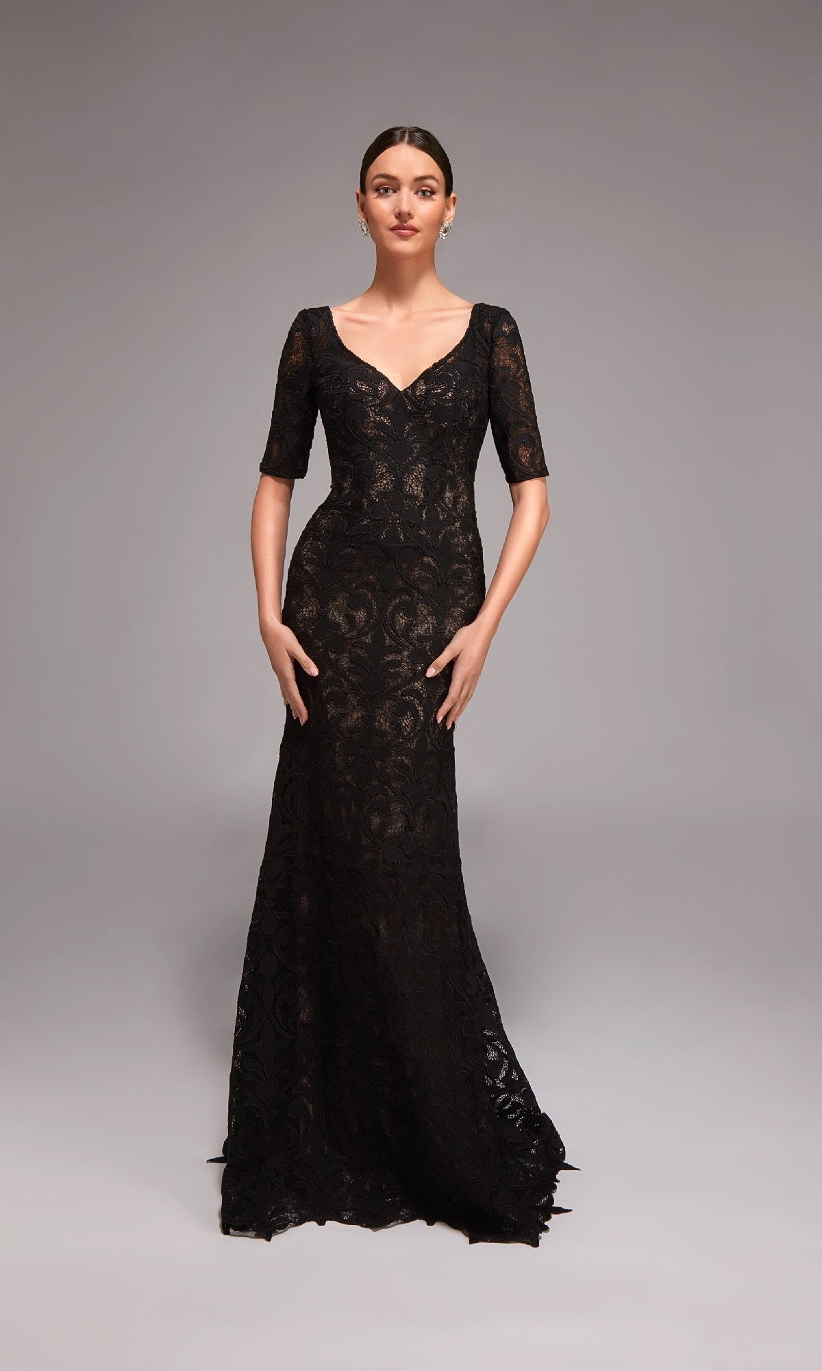 A woman poses in a studio wearing the Alyce 27871—a long formal dress featuring a fitted black lace overlay, short sleeves, and a V-neckline.